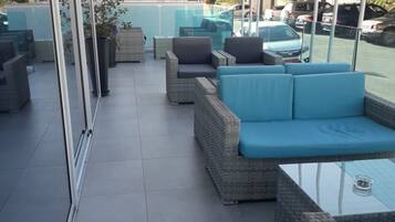 An outdoor patio area with wicker furniture, a glass table, and a view of parked cars and buildings in the background.