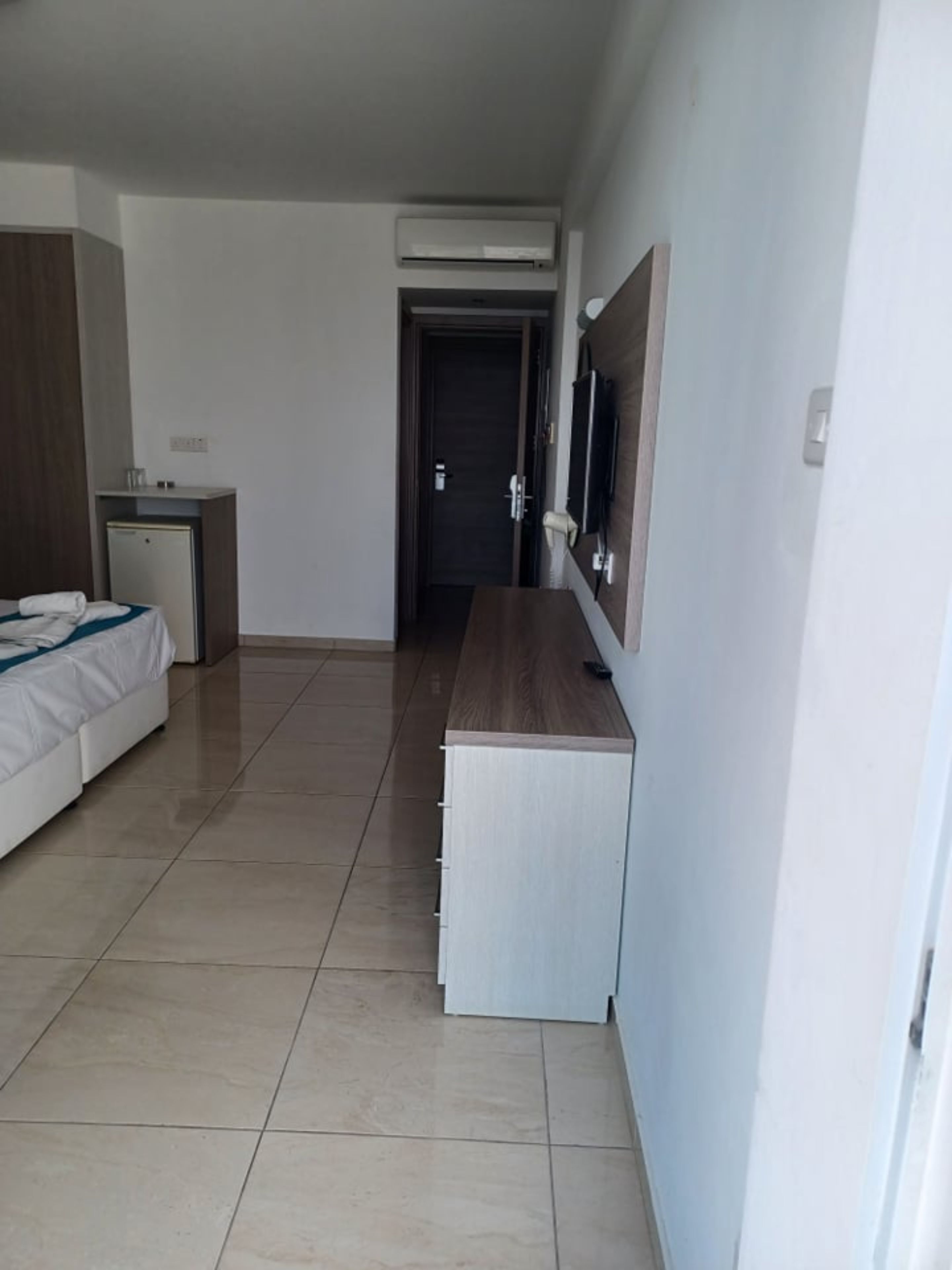 Quarto de hotel com uma cama, uma escrivaninha, um frigorífico pequeno, ar condicionado e uma televisão na parede.
