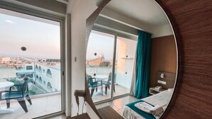 Une chambre d’hôtel dotée d’un grand miroir qui reflète un balcon avec des chaises et une table, un lit avec une tête de lit bleue et une vue sur la ville.