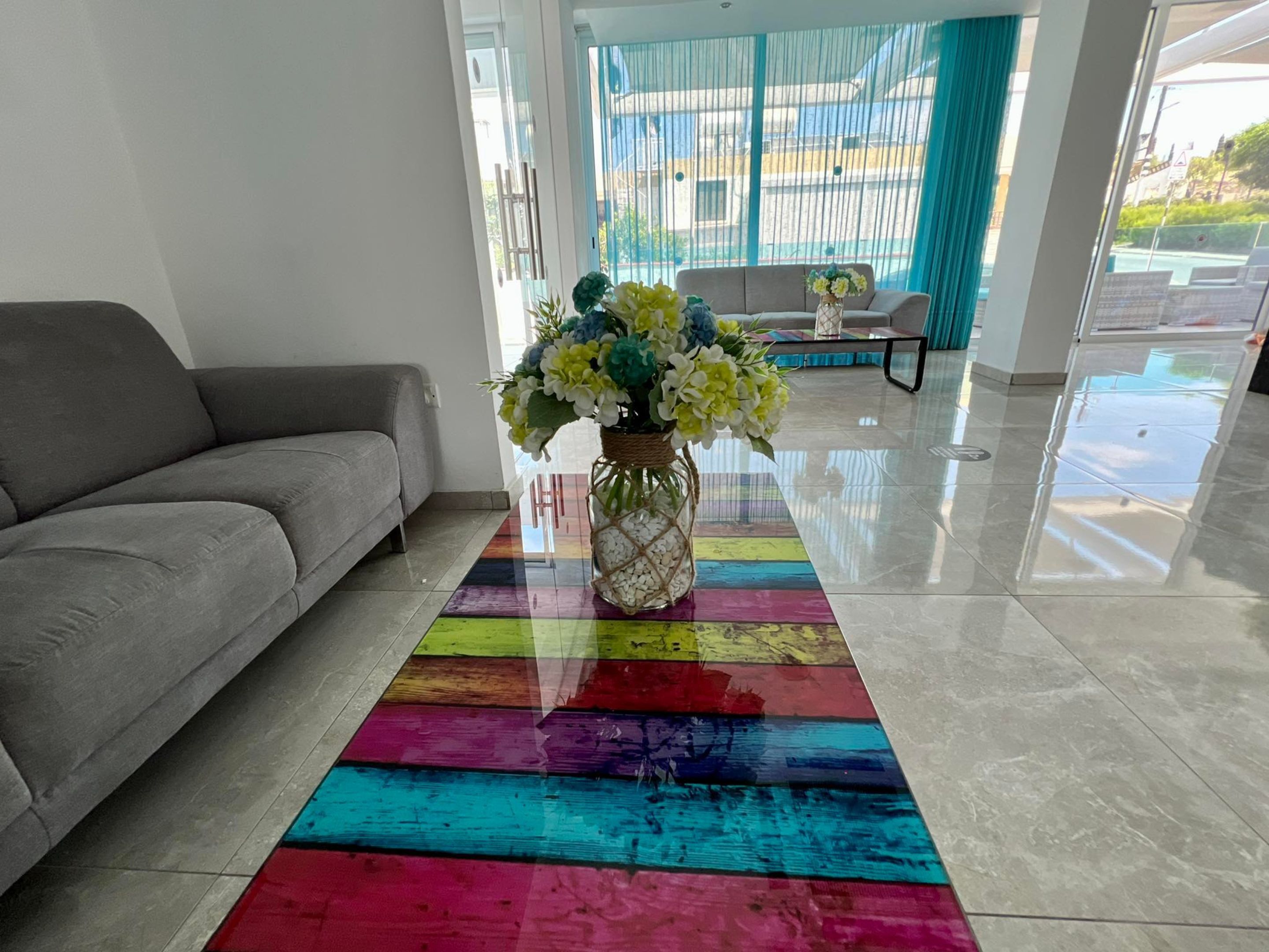 Un salon moderne avec un canapé gris, un tapis rayé aux couleurs vives et un vase de fleurs posé sur une table en verre.