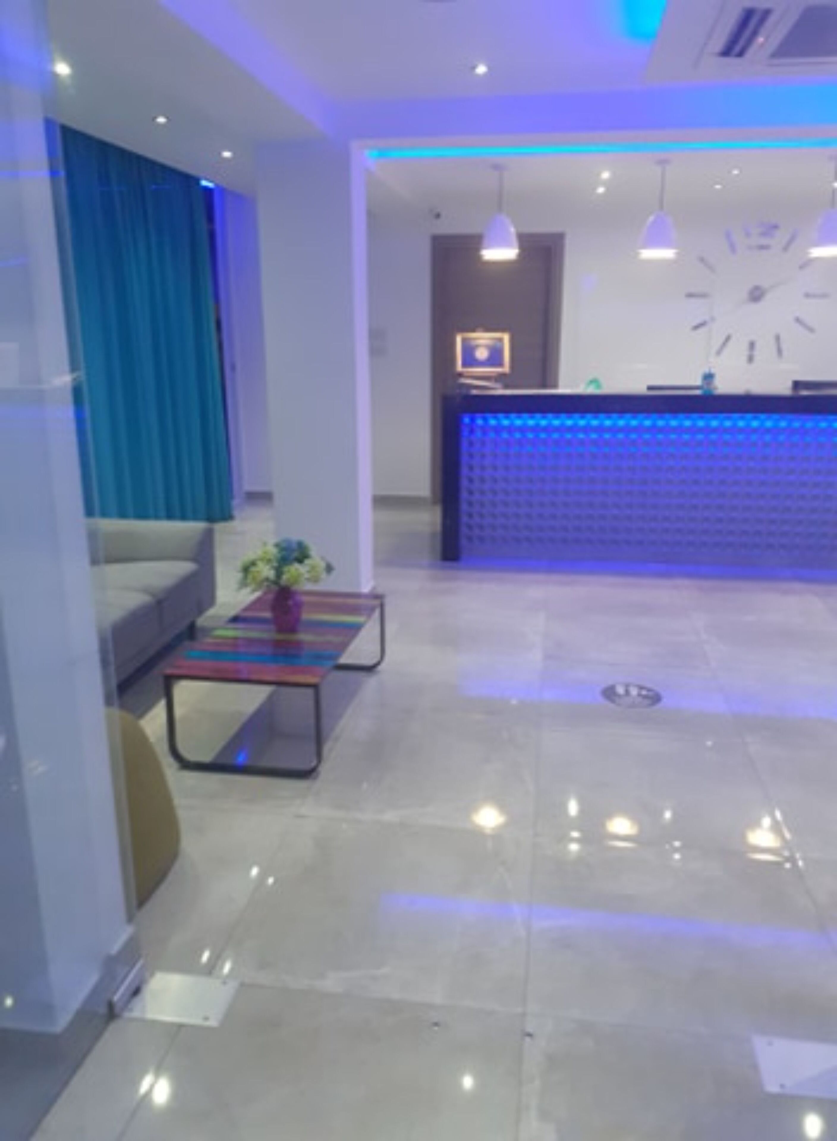 A modern hotel lobby with a reception desk, a sofa, a colorful coffee table, and a large clock on the wall.