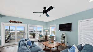 A spacious living room with a ceiling fan, a large TV mounted on the wall, and a view of the beach through the sliding glass doors. - Dog Friendly Private Beach House in Beautiful Fort Morgan, Alabama!  (Gulf Shores)