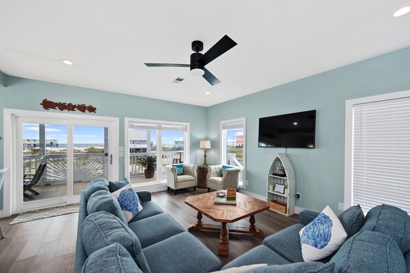 A spacious living room with a ceiling fan, a large TV mounted on the wall, and a view of the beach through the sliding glass doors.