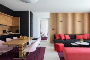 A modern living room with a wooden dining table, pink chairs, a black sofa with red cushions, and a red ottoman.