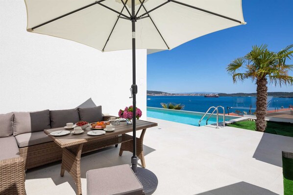 A terrace with a dining table, a sofa, and an umbrella. A pool and a view of the sea are visible in the background.