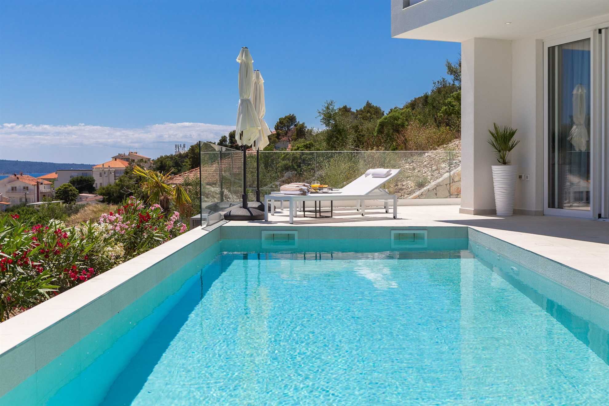 A swimming pool with lounge chairs and umbrellas, surrounded by a modern building and a view of the coastline.