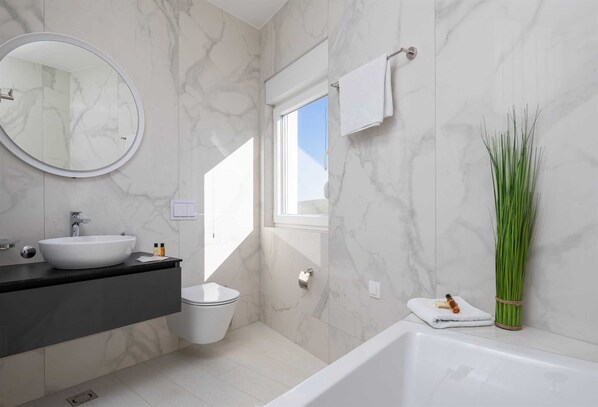 A modern bathroom with a white marble wall, a round mirror, a white sink, a toilet, and a bathtub. - Villa Adria View with Heated Pool II (Seget Donji)