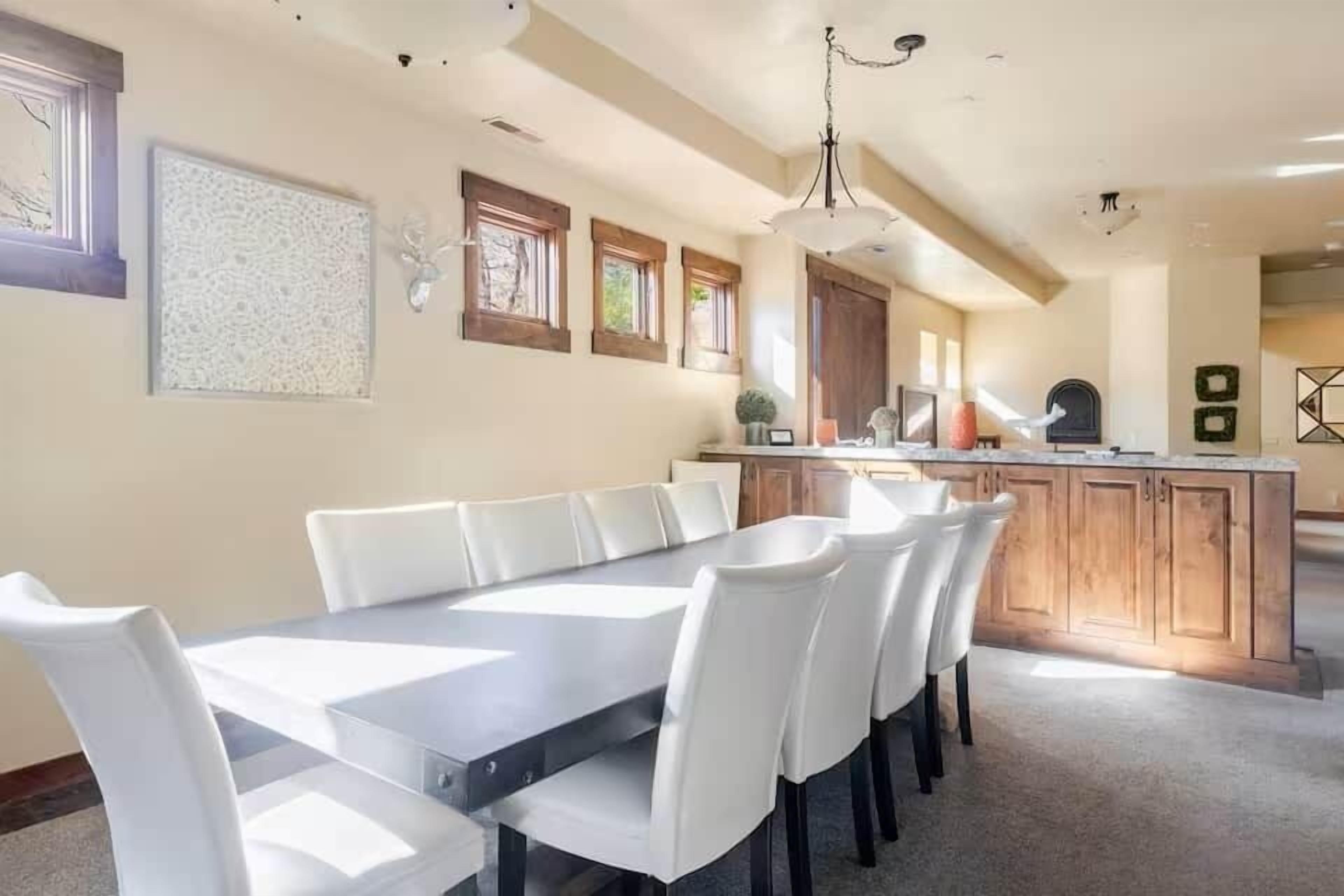 Un comedor con una mesa larga y sillas blancas, una cocina con armarios de madera y una lámpara colgante.