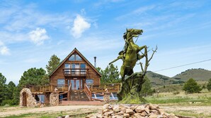 Eine Holzhütte mit einem Portal aus Stein, einer Holzdielen-Terrasse und einer Steinstatue eines Pferdes.
