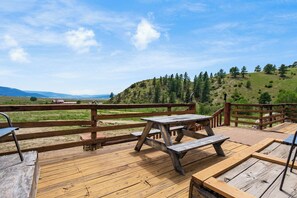 Une terrasse en bois, dotée de tables de pique-nique et d’une rambarde en bois, surplombe une colline verdoyante.