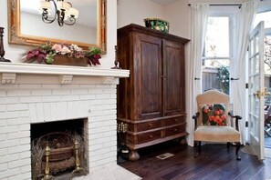 A room with a white brick fireplace, a large wooden cabinet, and a chair with a floral cushion.