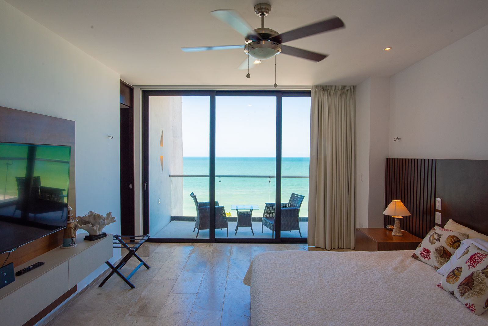 A hotel room with a large bed, a TV, a ceiling fan, a balcony with a view of the sea, and a table with chairs.