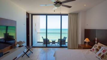 A hotel room with a large bed, a TV, a ceiling fan, a balcony with a view of the sea, and a table with chairs.
