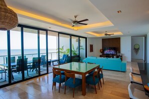 Ampla sala de estar com varanda vista para o mar, uma mesa de jantar com cadeiras, um sofá e uma TV.