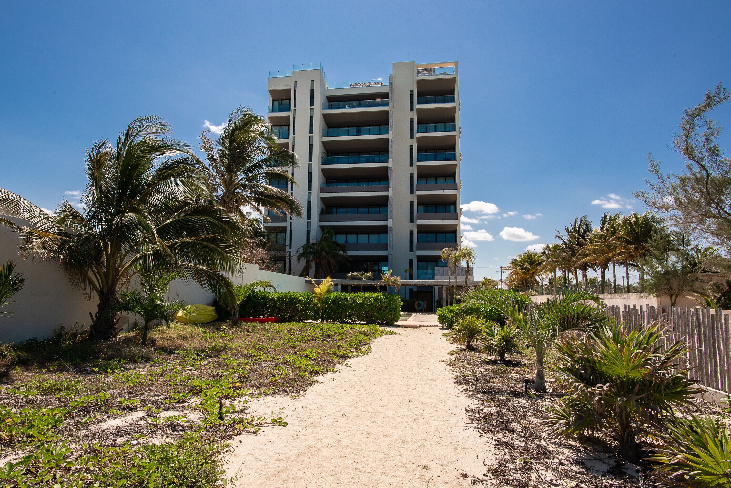 A multi-story hotel with a central entrance, surrounded by palm trees and a sandy pathway.