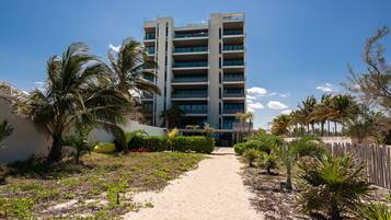 A multi-story hotel with a central entrance, surrounded by palm trees and a sandy pathway.
