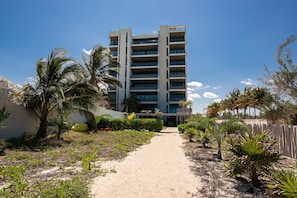 Hotel com vários andares, entrada central, rodeado por palmeiras e um caminho de areia.