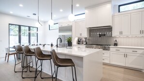 A modern kitchen with white cabinetry, a large island, bar stools, and a view of the outdoors. - Luxury New Build, Private Pool and Spa, Sleeps 10 all King, 4 min to Dt (Austin)