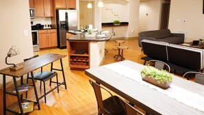 A modern kitchen with wooden cabinets, a stainless steel refrigerator, and a dining area with a wooden table and chairs. - The Tranquility Den (NRG,City Centre, Galleria) (Houston)