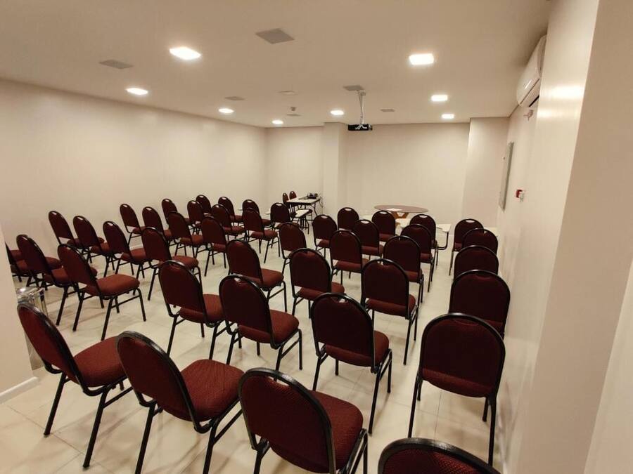 Un salón de conferencias con filas de sillas rojas, una pantalla de proyector y una mesa con sillas.