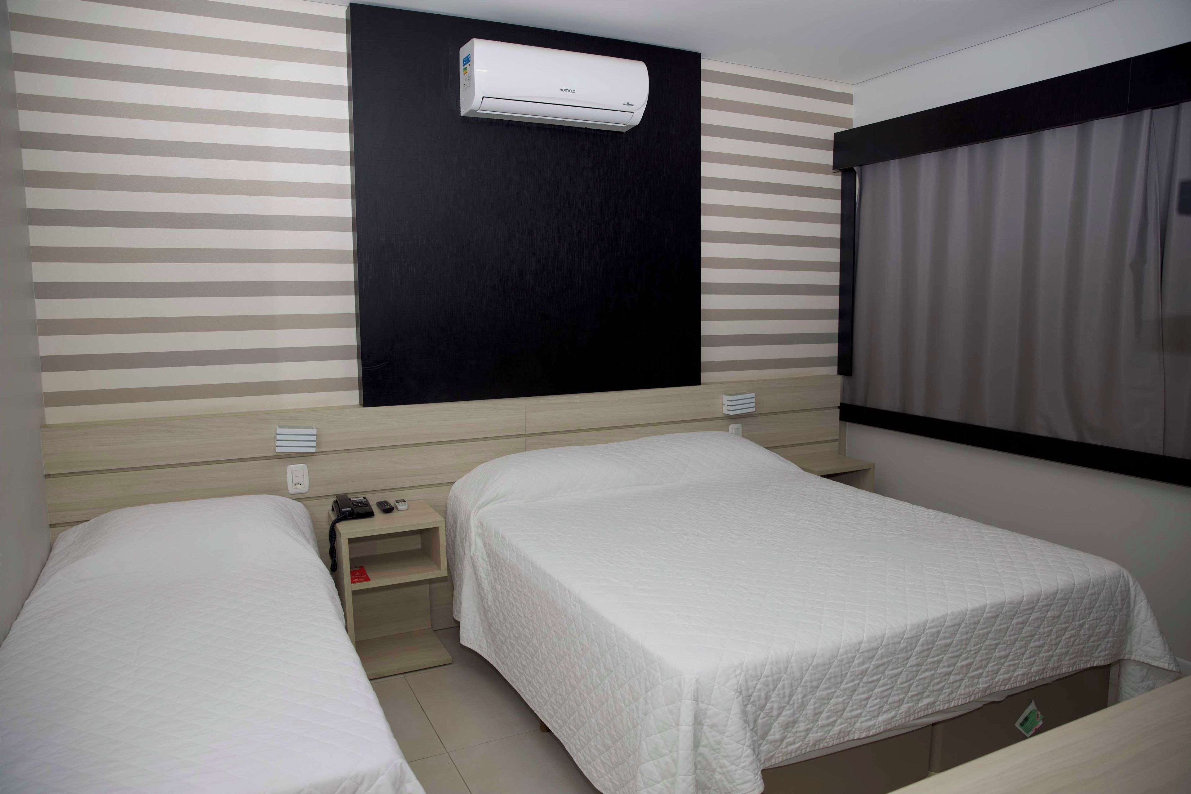 A hotel room with two single beds, a bedside table with a phone and a lamp, a wall-mounted air conditioner, and a window with curtains.