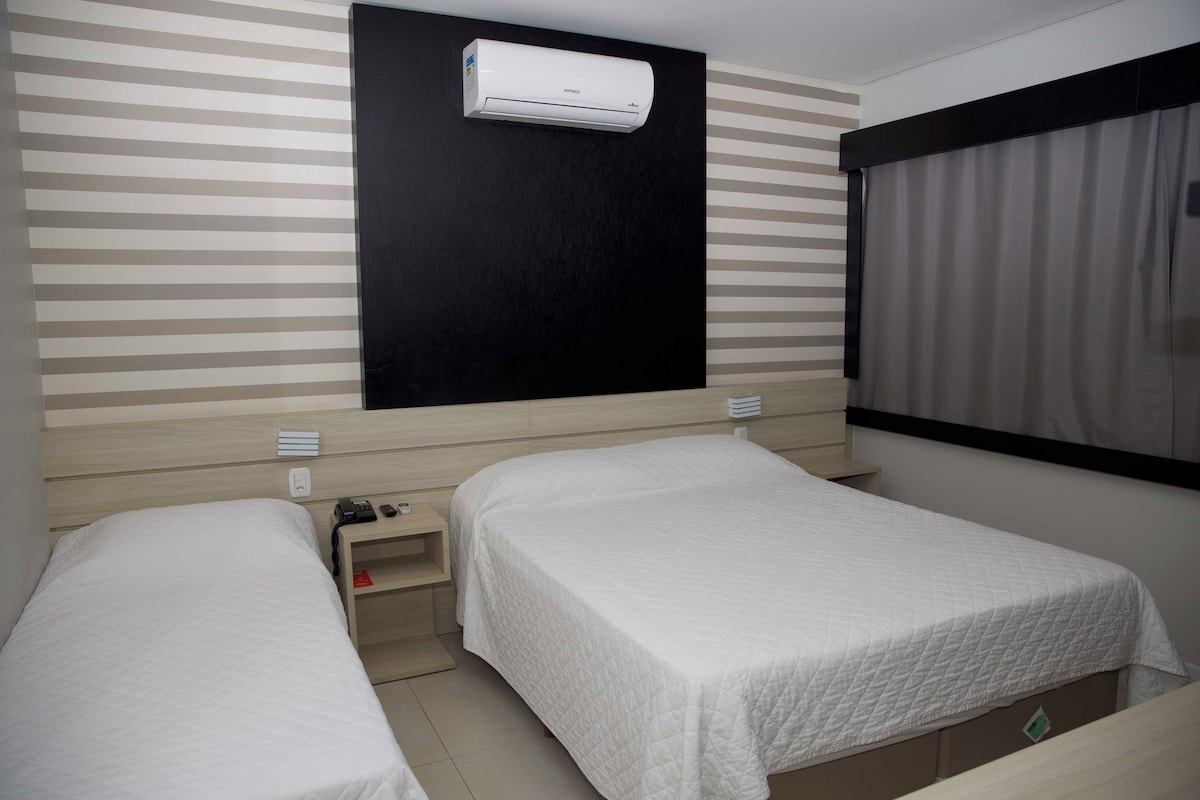 Una habitación de hotel con dos camas individuales, una mesita de noche con teléfono y lámpara, aire acondicionado instalado en la pared y una ventana con cortinas.
