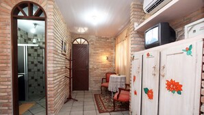 A compact living space with a tiled floor, a wooden door, a small table with a lace tablecloth, a television on a cabinet, and a patterned sofa. - Pousada Raja Canto do Mar (Ipojuca)