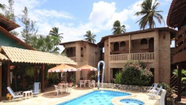 A swimming pool with lounge chairs and umbrellas, surrounded by buildings with balconies and palm trees. - Pousada Raja Canto do Mar (Ipojuca)