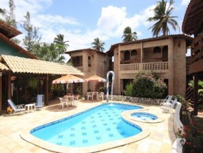 Piscina com espreguiçadeiras e guarda-sóis, cercada por edifícios com varandas e palmeiras.