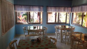 A dining area with two tables and chairs, large windows with curtains, and a view of greenery outside. - Pousada Raja Canto do Mar (Ipojuca)