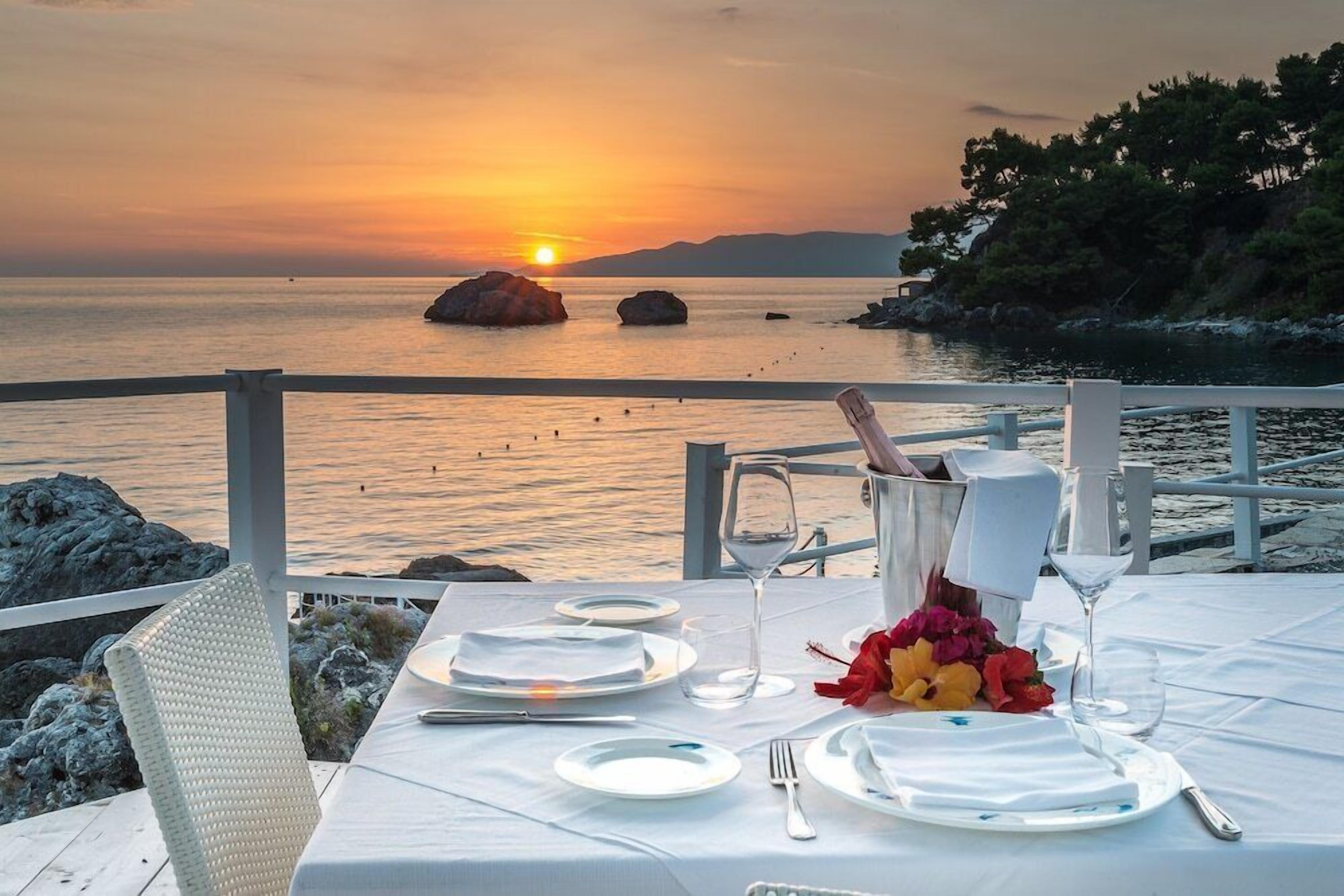 Mesa para dos con vista al atardecer, mantel blanco, platos, cubiertos y un jarrón de flores.