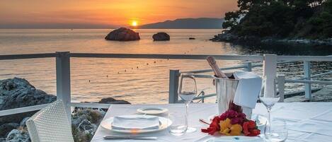 Mesa para dos con vista al atardecer, mantel blanco, platos, cubiertos y un jarrón de flores.