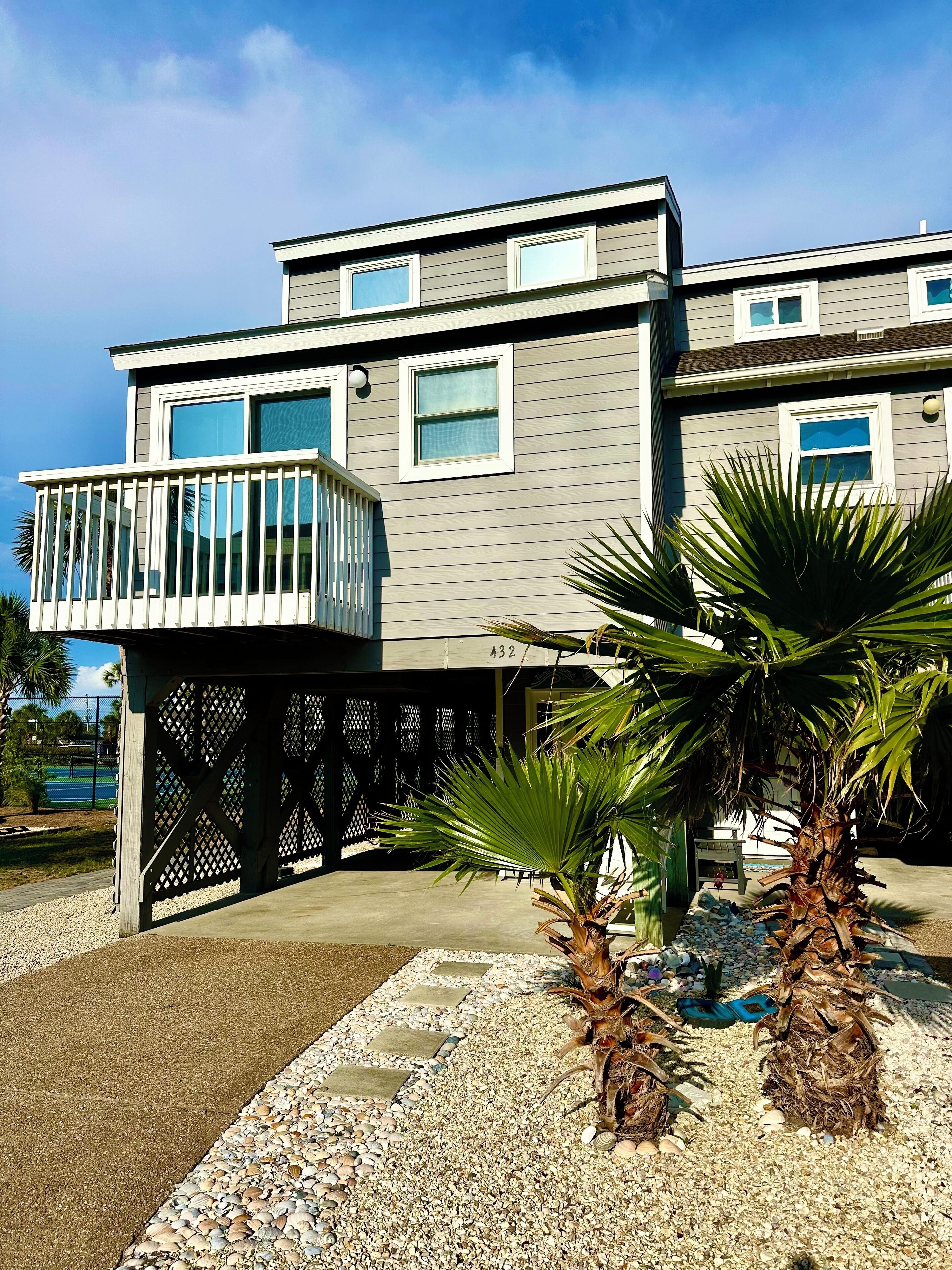 A modern two-story house with a balcony, palm trees, and a tennis court in the background.