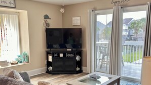 Un salon doté d’un ventilateur de plafond, d’un téléviseur à écran plat, d’un meuble de rangement noir portant l’inscription « Beach » et d’une table basse en verre.