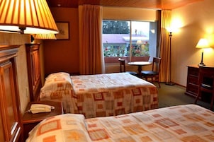 Quarto de hotel com duas camas, uma mesinha, uma cadeira e uma janela com cortinas.