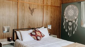 Une chambre à coucher aux murs lambrés, avec un lit, une table de chevet, un attrape-rêves fixé au mur et un ventilateur de plafond.