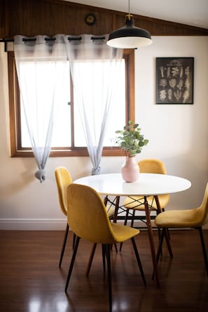 A dining area with a round table, yellow chairs, a pink vase with greenery, and a pendant light.