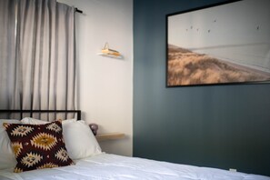 A bedroom with a bed, a pillow, a wall-mounted shelf, a framed picture, and a curtain.