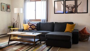 Un salon avec un canapé d’angle gris, une table basse en bois, un lampadaire et un tapis coloré. Trois tableaux encadrés ornent le mur, et une fenêtre est agrémentée de rideaux.