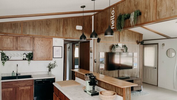 Une cuisine moderne avec des meubles en bois, un îlot central, des luminaires suspendus, un four encastré et un grand téléviseur.