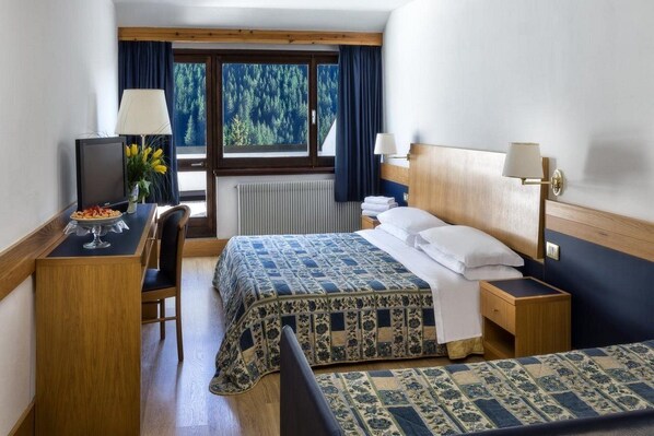 A hotel room with a bed, a desk with a computer, a chair, a window with a view of trees, and a lamp on the bedside table. - Hotel Ski Club Regina E Fassa (Mazzin)