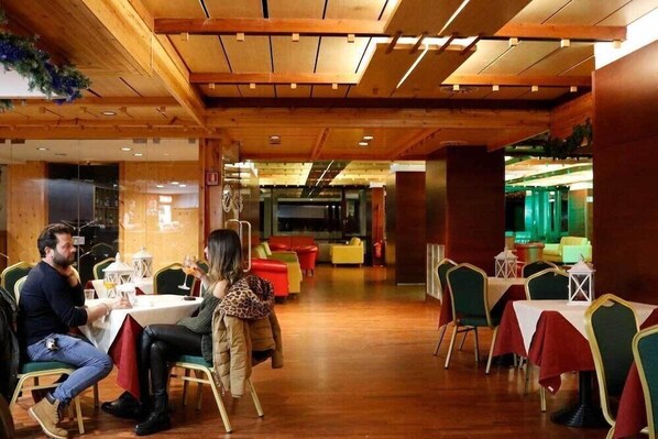 A restaurant with wooden ceiling beams, tables with red tablecloths, and green chairs. - Hotel Ski Club Regina E Fassa (Mazzin)