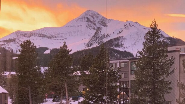 A mountain with snow-covered peaks and a sunset sky. There are buildings and trees in the foreground. - Studio getaway steps away from Big Sky Resort (Big Sky)