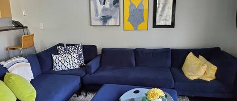 Un salon avec un canapé d’angle bleu, une petite table blanche ornée d’un vase de fleurs et trois œuvres d’art encadrées accrochées au mur.