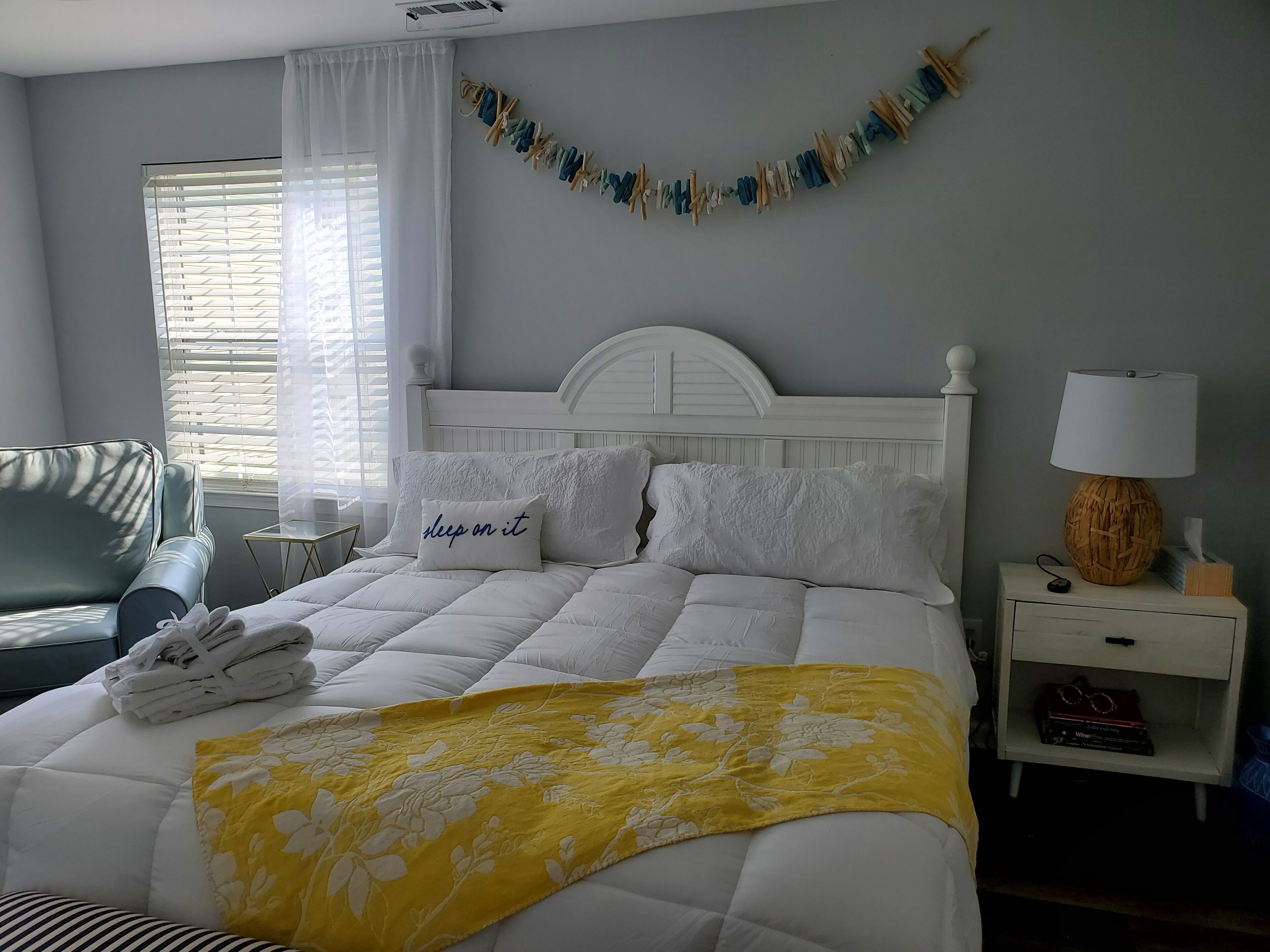 Een netjes opgemaakt bed met een bloemenkleed, een kussen met de tekst 'slaap erover' en een nachtkastje met een lamp en een boek.