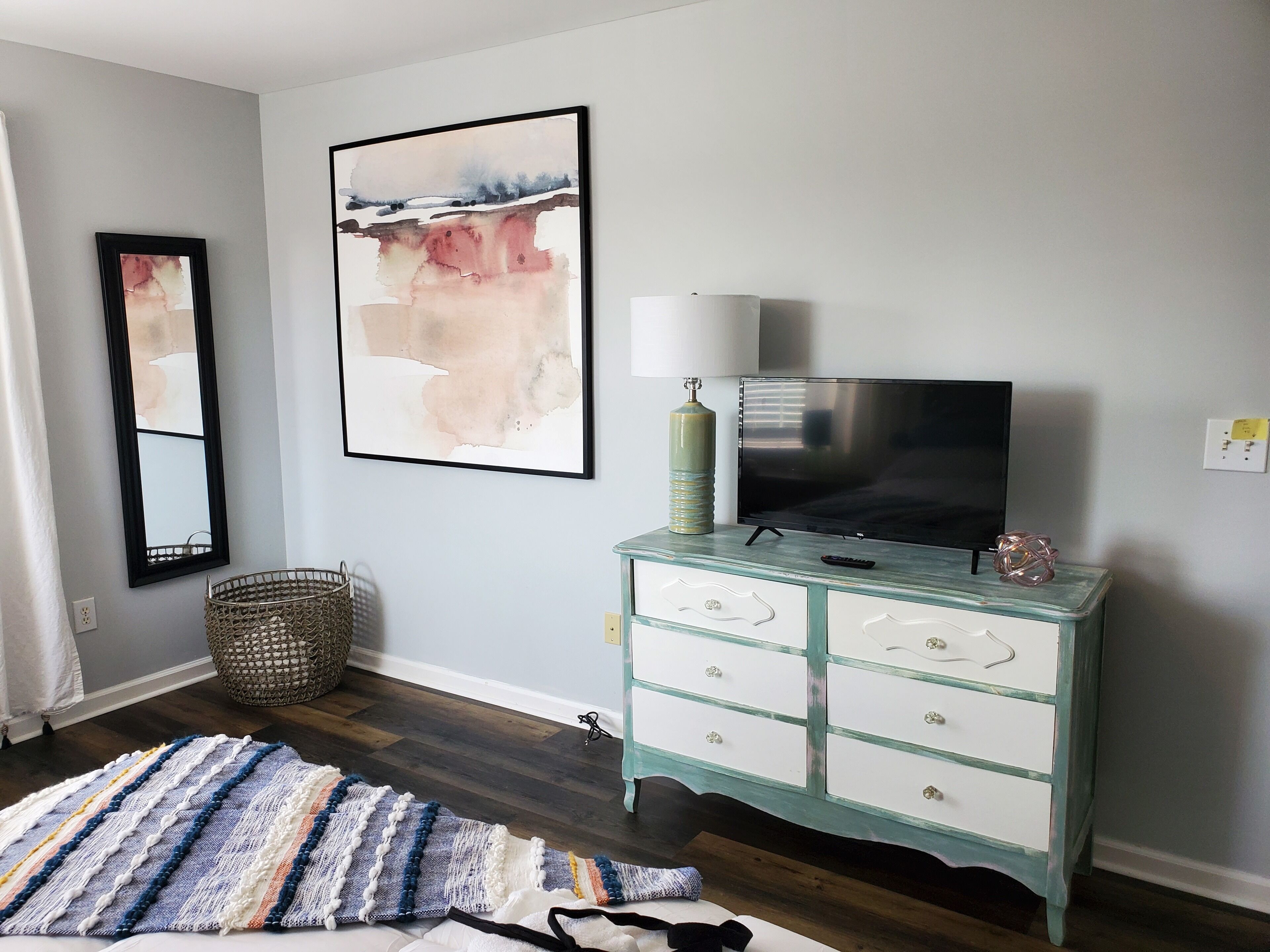 A room with a teal dresser, a large abstract painting, a mirror, and a television on a stand.