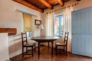 A dining area with a round table and two chairs, a lamp, a window with curtains, and a blue door.