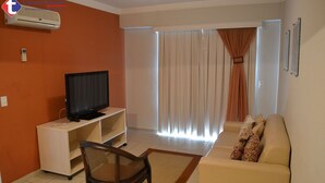 Una sala de estar con televisor sobre un mueble bajo, un sofá, un sillón y un ventanal con cortinas.
