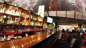 Una zona de bar con una barra larga, estantes llenos de botellas, vasos y un área de asientos con sillones acolchados.