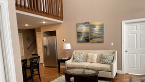 A living room with a beige sofa, patterned rug, and a glass coffee table. A kitchen area with a refrigerator and a dining table is visible in the background. - Lake House with stunning view of Lake Cumberland (Somerset)